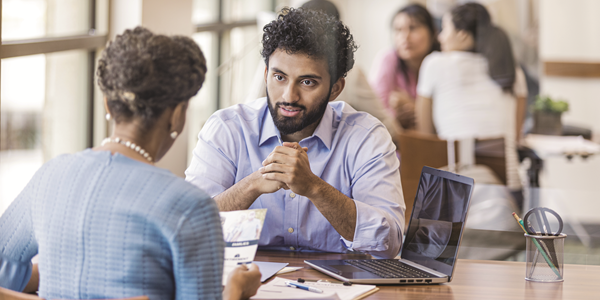 Image of two colleagues speaking to each other at a table. A pile of papers and a computer are on the table.